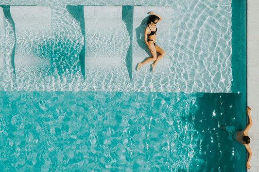 Vrouw liggend in een helderblauw zwembad, gezien van bovenaf, met zonlicht dat speelse patronen op het water tekent.