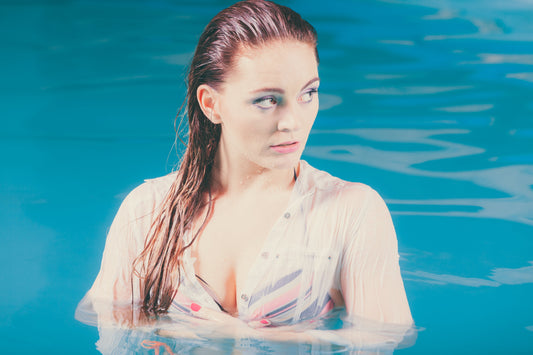 Jonge vrouw in helderblauw water, natte haren op haar schouders, blik opzij gericht, kwetsbaar en bedachtzaam in zomers licht.