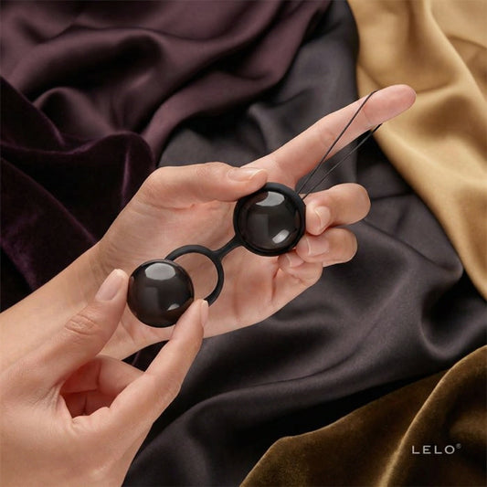 Twee handen houden elegante zwarte LELO benwa-balletjes vast tegen een donkere achtergrond, voor bekkenbodemtraining
