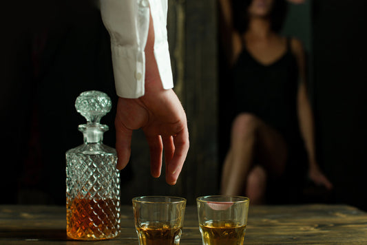 Kristallen karaf en twee glazen op een tafel, met op de achtergrond een vrouw in het halfduister die afwachtend zit.