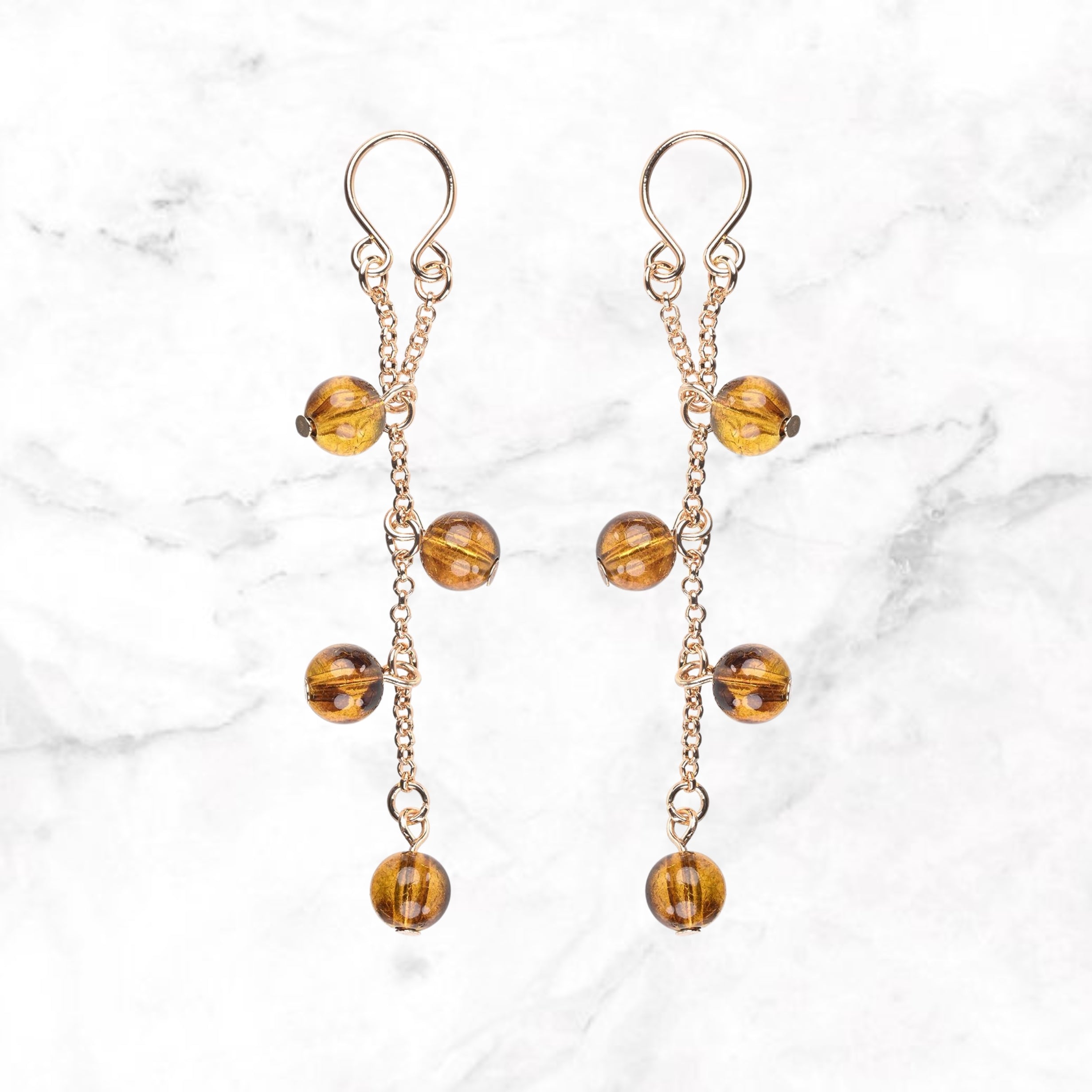Twee tepelsieraden met goudkleurige kettinkjes en amberkleurige bolkralen op marmer