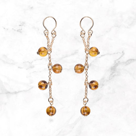 Twee tepelsieraden met goudkleurige kettinkjes en amberkleurige bolkralen op marmer